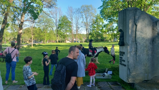 Zirgu izjādes aploks muzeja pļavā. Bērni un vecāki gaida izjādes rindā. Zemnieku saimniecības "Kurģi"  zirgi aplokā. Uz Tinker šķirnes zirga, meitenīte izjādē.  Pavadone pieved poniju uz izjādes sākumu vietu.