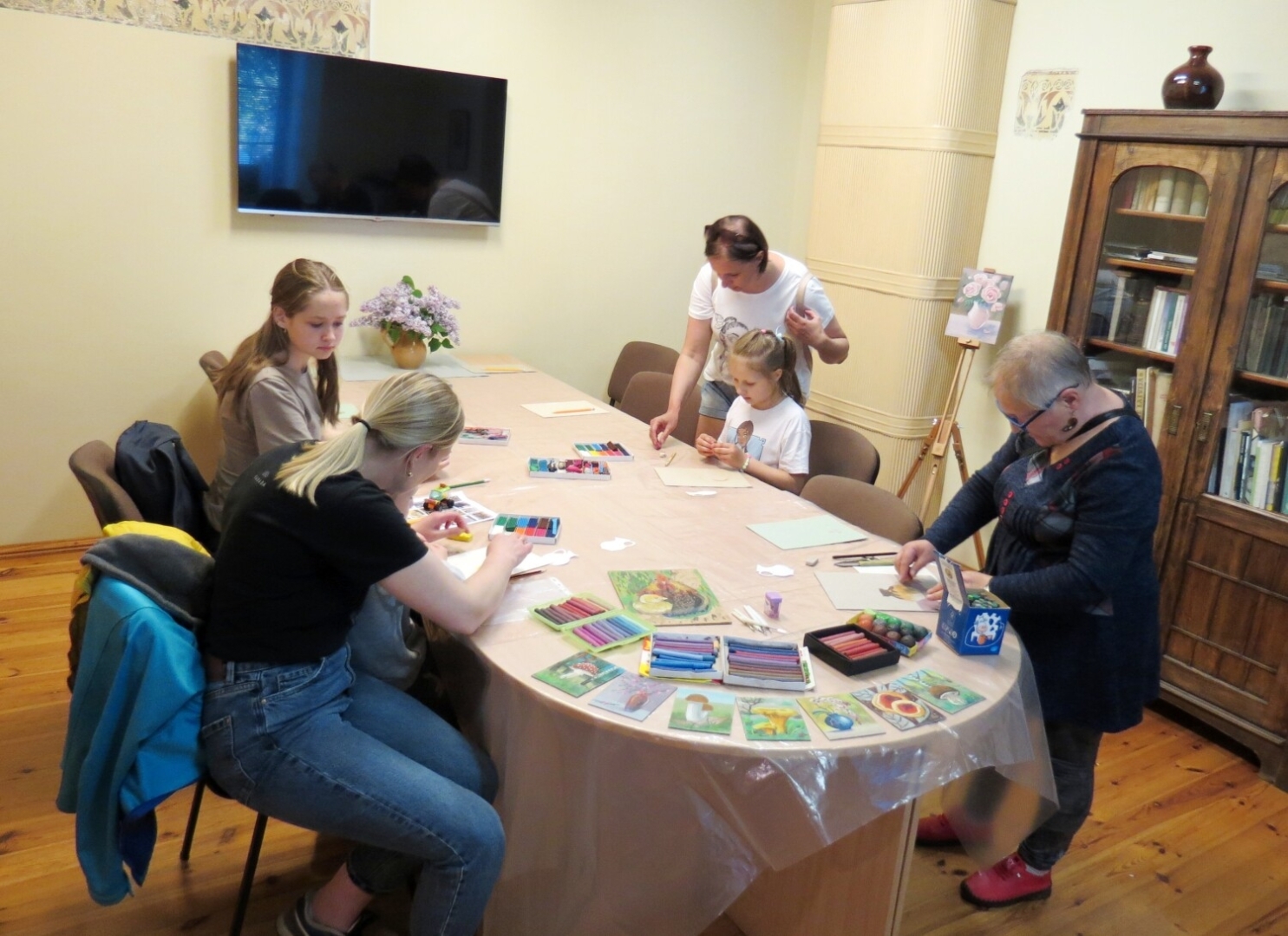 Priekšplānā kreisajā pusē jaunietes ar plastilīna gleznu darbu pamatnēm. Labajā pusē Vija Meistare Muzeju nakts akcijas laikā, iesākot plastilīna gleznu darbnīcu Valkas novadpēniecības muzeja apmeklētāju telpā. Aizmugurē meitenīte ar piegaušo pie sava darba pamatnītnes. 